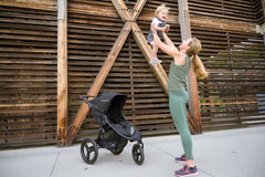 Mother holding child in air next to Bumbleride Australia Speed Jogging Stroller in Black on sidewalk with wood wall in background