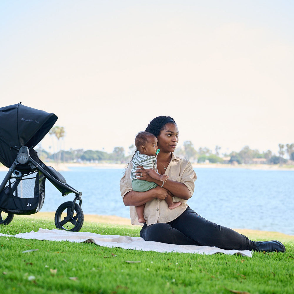 
                      
                        Mother holding child to chest sitting on white blanket on grass in front of the bay with Bumbleride Indie in Black parked next to them- New Collection 2022 - Global
                      
                    