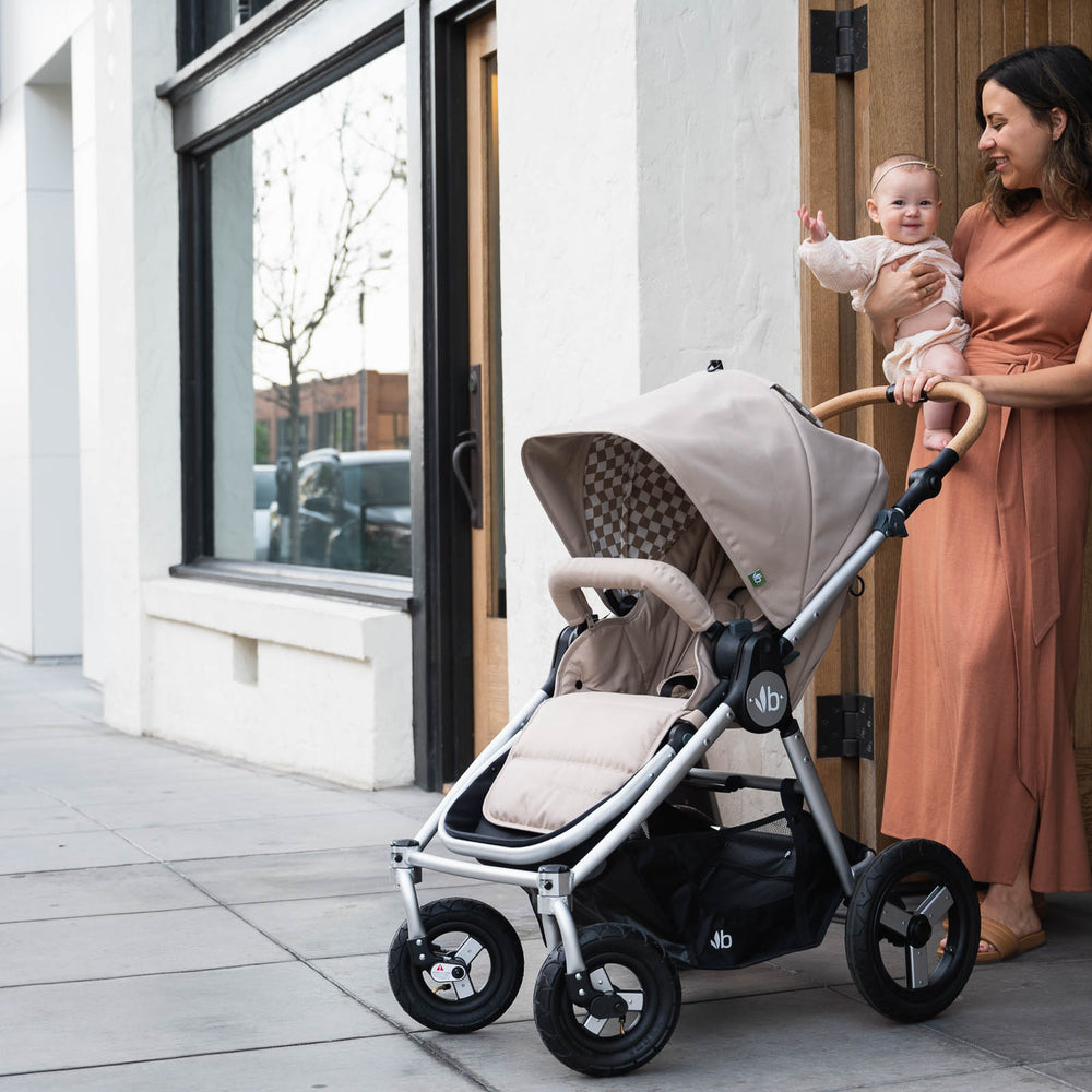 
                      
                        Picture of mother holding child to chest while standing in front of wood door with Bumbleride Era reversible stroller in Sand in front of them. New Collection 2022. Global
                      
                    