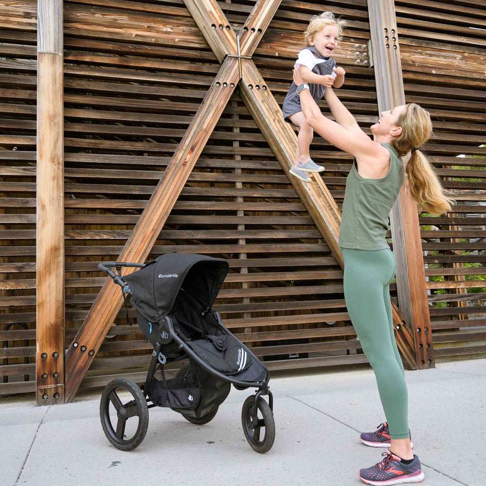 
                      
                        Mother holding child in air next to Bumbleride Australia Speed Jogging Stroller in Black on sidewalk with wood wall in background
                      
                    