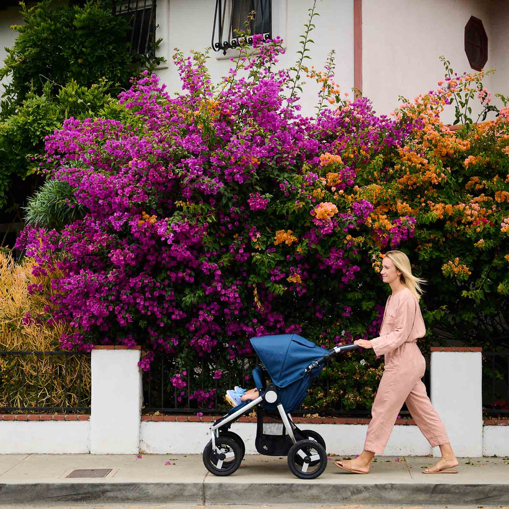 
                      
                        Mother pushing Bumbleride Indie Twin double stroller in Maritime in front of bright colored flowers on sidewalk. Global
                      
                    